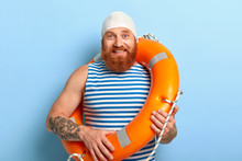 Summer Vacation Concept. Cheerful Man With Ginger Beard, Wears Swimcap And Sailor T Shirt, Poses With Lifering, Works As Lifeguard At Beach, Has Fun. Accident Prevention And Swimming Equipment