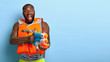Indoor shot of emotional dark skinned man has fight with someone, plays with water gun, stares into distance with widely opened mouth, enjoys real battle, wears protective lifevest. Summer activity