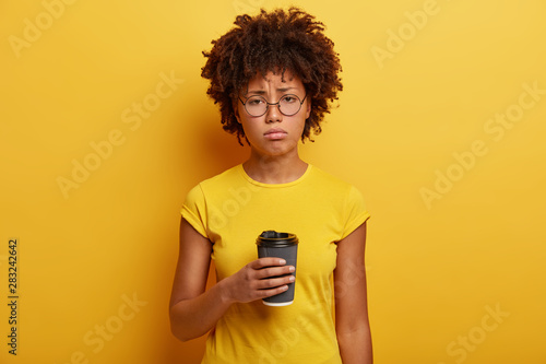 Unhappy dark skinned student holds disposable cup of caffeine beverage, has hard Monday, wears round glasses and t shirt, stands over yellow wall, being in low spirit. Youth and drinking concept
