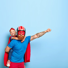 Powerful Dad Gives Piggy Back To Child, Demonstrates Courage, Makes Flying Gesture, Wears Helmet, Red Mask, Cape, Have Fun Together, Stand Against Blue Wall With Empty Space, Brave Help Someone Else