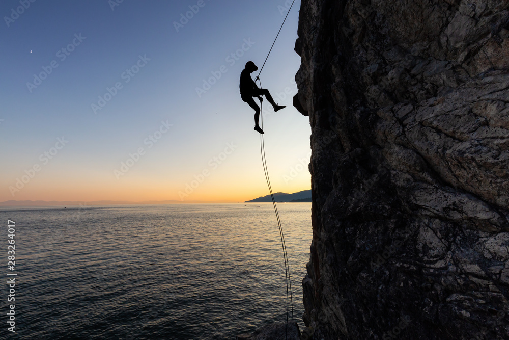 Silhouette of a Unrecognizable man rappelling down a steep cliff on the rocly ocean coast during a sunny summer sunset. Taken in Lighthouse Park, West Vancouver, British Columbia, Canada. - obrazy, fototapety, plakaty 