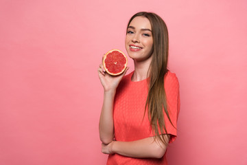 attractive smiling young woman holding cut grapefruit and looking at camera on pink