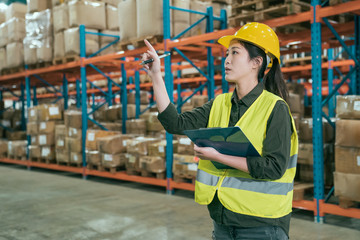 attractive woman in warehouse checking inventory levels of goods on shelf. lady worker in hard hat and safety vest point finger looking up counting parcels and cardboard boxes in large storehouse.