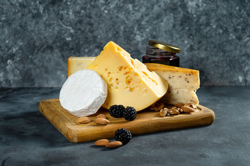 Cheese Assortment on a dark stone background with copy space. Different types: Camembert, cheese with spices, Dutch cheese on wooden cutting board. almond, blackberries and jam with cheese. Soft focus