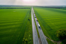 Convoys With Cargo. Trucks On The Higthway Sunset. Cargo Delivery Driving On Asphalt Road Along The Green Fields. Seen From The Air. Aerial View Landscape. Drone Photography.