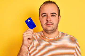 Sticker - Young man holding blue credit card standing over isolated yellow background with a confident expression on smart face thinking serious