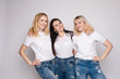 © Вячеслав Косько - Side view of three beautiful young women standing one by one. Blonde and brunette girls in white t-shirts and jeans posing at camera. Happy friends in casual clothes smiling being close to each other.