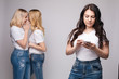 © Вячеслав Косько - Front view of offended brunette with folded arms looking aside while two blondes standing behind and gossiping. Female friends laughing and talking on grey isolated background. Concept of sneering.