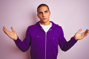 Sticker - Young handsome man wearing purple sweatshirt over pink isolated background clueless and confused expression with arms and hands raised. Doubt concept.