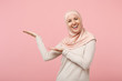Cheerful smiling young arabian muslim woman in hijab light clothes posing isolated on pink background in studio. People religious Islam lifestyle concept. Mock up copy space. Pointing hands aside.
