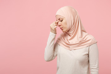 Poster - Tired arabian muslim woman in hijab light clothes posing isolated on pink background in studio. People religious Islam lifestyle concept. Mock up copy space. Keeping eyes closed, putting hand on nose.