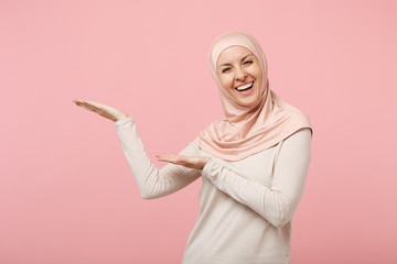 Cheerful smiling young arabian muslim woman in hijab light clothes posing isolated on pink background in studio. People religious Islam lifestyle concept. Mock up copy space. Pointing hands aside.