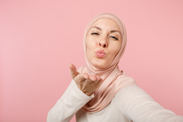 Close up arabian muslim woman in hijab light clothes posing isolated on pink background. People religious lifestyle concept. Mock up copy space. Doing selfie shot on mobile phone, sending air kiss.