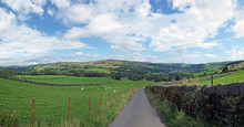 A Wide Panoramic View Of Yorkshire Dales Countryside With A Narrow Country Lane With Stone Walls And Fences Around Grass Meadows With Grazing Sheep And Pennine Villages Amoung Hillside Trees
