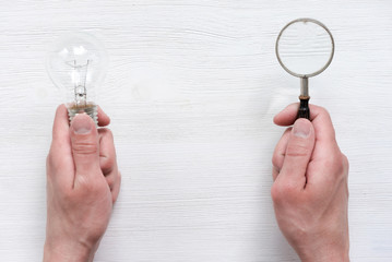 Light bulb and magnifying glass in hands on a white wooden background with a copy space.