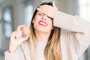 Poster - Young beautiful woman holding fresh egg at home stressed with hand on head, shocked with shame and surprise face, angry and frustrated. Fear and upset for mistake.