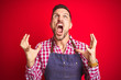 © Krakenimages.com - Young handsome man wearing shop owner apron uniform over red isolated background crazy and mad shouting and yelling with aggressive expression and arms raised. Frustration concept.