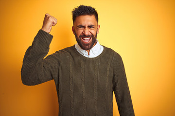 Canvas Print - Young indian man wearing green sweater and shirt standing over isolated yellow background angry and mad raising fist frustrated and furious while shouting with anger. Rage and aggressive concept.