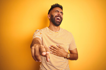 Sticker - Young indian man wearing t-shirt standing over isolated yellow background laughing at you, pointing finger to the camera with hand over body, shame expression