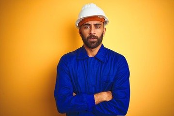 Wall Mural - Handsome indian worker man wearing uniform and helmet over isolated yellow background skeptic and nervous, disapproving expression on face with crossed arms. Negative person.