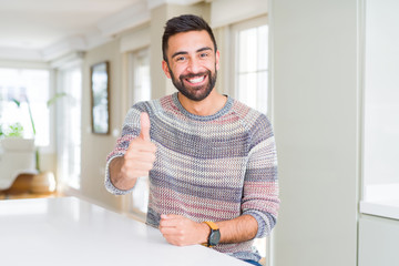 Handsome hispanic man wearing casual sweater at home doing happy thumbs up gesture with hand. Approving expression looking at the camera with showing success.