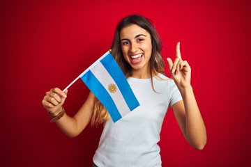 Canvas Print - Young beautiful woman holding argentine flag over red isolated background surprised with an idea or question pointing finger with happy face, number one