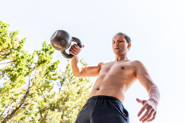 Young shirtless man exercising with kettlebell outdoors outside park backyard doing rack and lift exercise low angle view with sky
