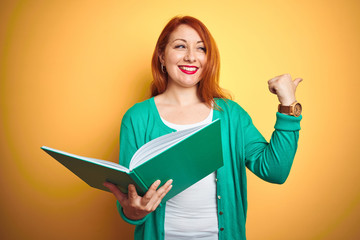 Poster - Young redhead student woman reading green book over yellow isolated background pointing and showing with thumb up to the side with happy face smiling