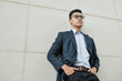 © DragonImages - Confident young Vietnamese businessman in glasses putting hands in pockets when standing at building wall and looking in front of him