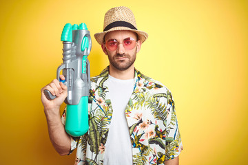 Sticker - Young man wearing hawaiian flowers shirt holding water gun over yellow isolated background with a confident expression on smart face thinking serious