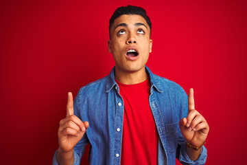 Young brazilian man wearing denim shirt standing over isolated red background amazed and surprised looking up and pointing with fingers and raised arms.