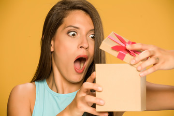 Young shocked woman opening a present box on yellow background