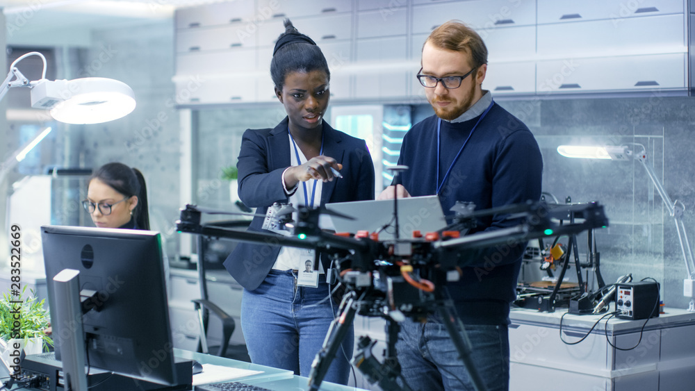 Caucasian Male and Black Female Engineers Working on a Drone Project with Help of Laptop and Taking Notes. He Works in a Bright Modern High-Tech Laboratory. - obrazy, fototapety, plakaty 