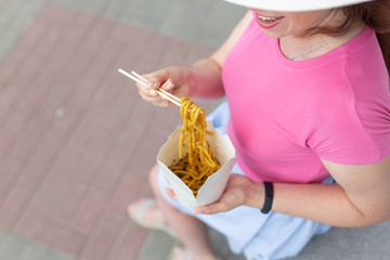 Beautiful young unidentified woman in a hat eats Chinese noodles while walking in the park on a warm summer day. Takeaway food concept.