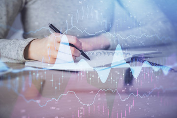 Hands writing information about stock market in notepad. Forex chart holograms in front. Concept of research. Multi exposure