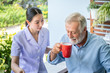 Nurse assist elderly senior man to drink coffee with mug in hand at nursing home