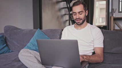 Poster - Smiling handsome bearded man using laptop computer while sitting on sofa at home