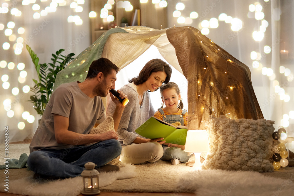 family, hygge and people concept - happy mother, father and little daughter reading book with torch light in kids tent at night at home - obrazy, fototapety, plakaty 