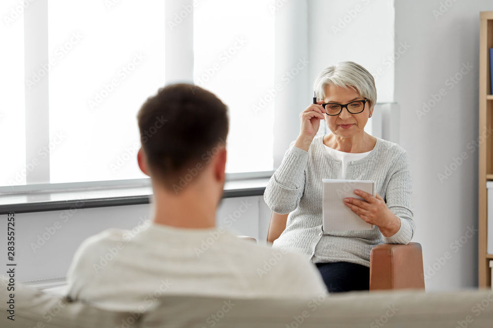 psychology, mental therapy and people concept - senior woman psychologist with clipboard and young man patient at psychotherapy session - obrazy, fototapety, plakaty 