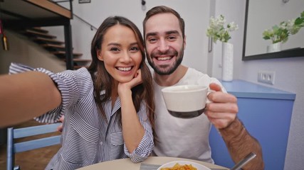 Wall Mural - Happy lovely couple having breakfast and making selfie while sitting together on kitchen