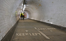 Cyclist Wearing Yellow Jacket Walking With His Bicycle Inside Greenwich Foot Tunnel Under River Thames, More Blurred Pedestrians In Background, Keep Left Sign On Floor