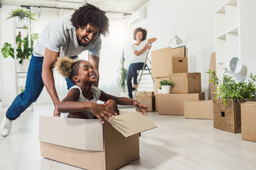 Happy playful  African American family moving in new apartment, little preschooler daughter sitting in cardboard boxes, father rolling her,  purchase property concept