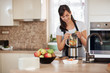 © nenadaksic - Beautiful Caucasian smiling woman in apron putting vegetables and fruits in blender while standing in kitchen.