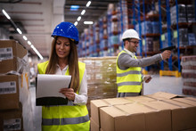 Female Factory Worker In Reflective Uniform With Hardhat Helmet Checking New Arrival Of Goods In Warehouse While Worker Using Bar Code Reader In Background. Logistics And Distribution.