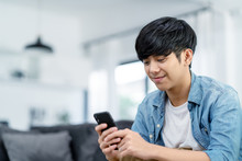 Happy Asian Teenager Using Smart Phone And Smiling On Sofa Living Room At Home. Asian Man Holding And Using Cellphone For Searching Data And Social Medie On Internet. Teenager And Technology Concept.