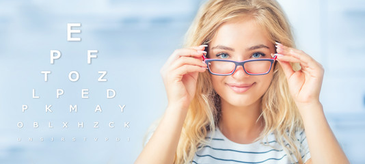 Wall Mural - Portrait of young woman holding glasses and looking at camera. Ophthalmological concept with eye test chart