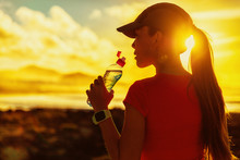 Fitness Woman Drinking Water From Sports Bottle On Afternoon Workout After Run Training Jogging Outdoors At Sunset. Girl Wearing Running Cap Silhouette Against Sun Flare.
