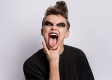 Crazy Teen Boy With Spooking Make-up Making Rock Gesture On Gray Background. Teenager In Style Of Punk Goth Dressed In Black Shows Tongue And Doing Heavy Metal Rock Sign. Problems Of Transitional Age