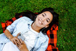 © Marharyta Hanhalo - Music application on smartphones and cell mobile phone in hands with earphones. Young asian woman listening to music and relax on a green grass in park. Happy lifestyle people. Podcast online