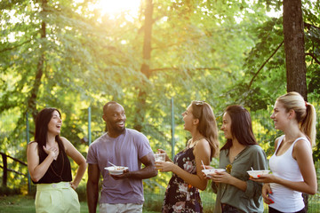 Wall Mural - Group of happy friends eating and drinking beers at barbecue dinner on sunset time. Having meal together outdoor in a forest glade. Celebrating and relaxing. Summer lifestyle, food, friendship concept